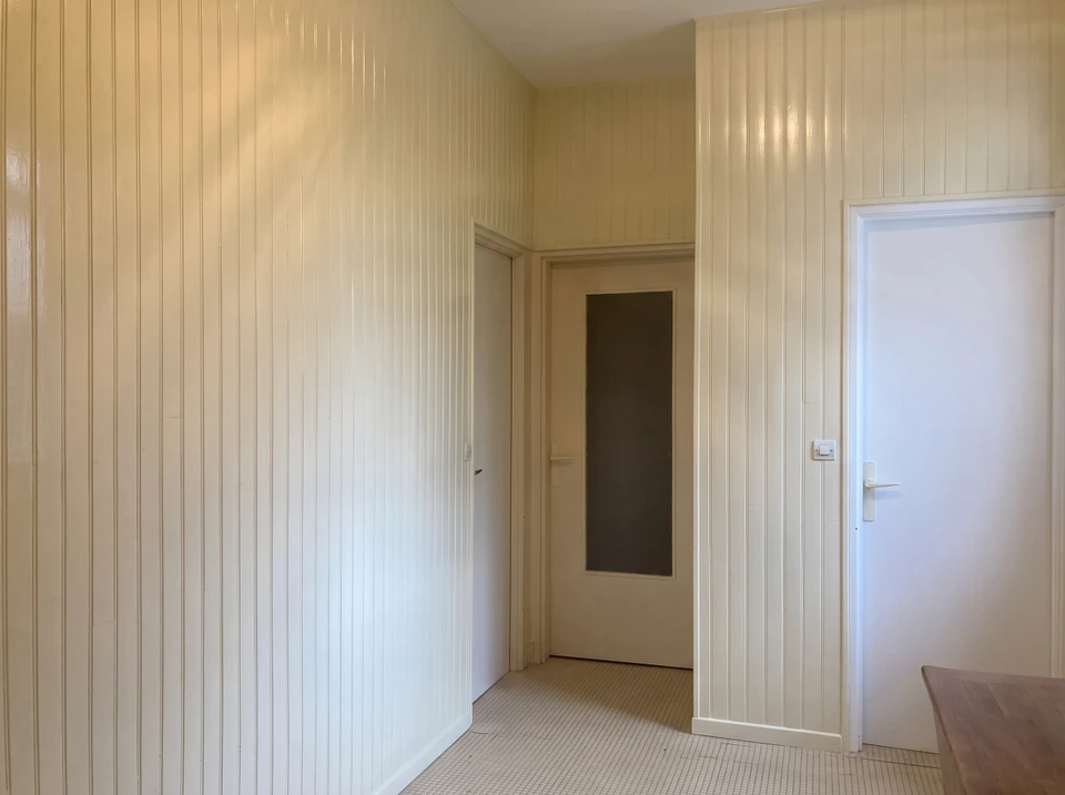 Deux ouvriers en combinaisons bleue et verte travaillent à genoux sur un chantier de rénovation intérieure, manipulant des outils de mesure près d'un mur blanc